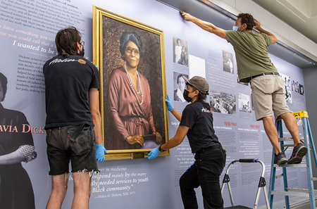 A man and a woman dressed in matching black T-shirts lift the portrait of Helen O. Dickens into place within an expanded exhibit housing, while a man on a stepladder does work on the display backdrop.